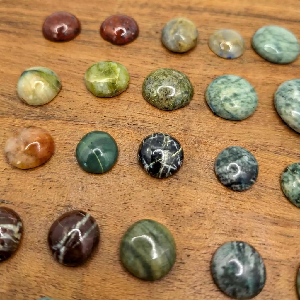 A varied collection of small cabochon gemstones arranged on a polished wood surface. Green, brown, blue, and cream stones show unique banding, orbicular patterns, and natural mineral inclusions. A quarter at the edge of the frame provides scale.