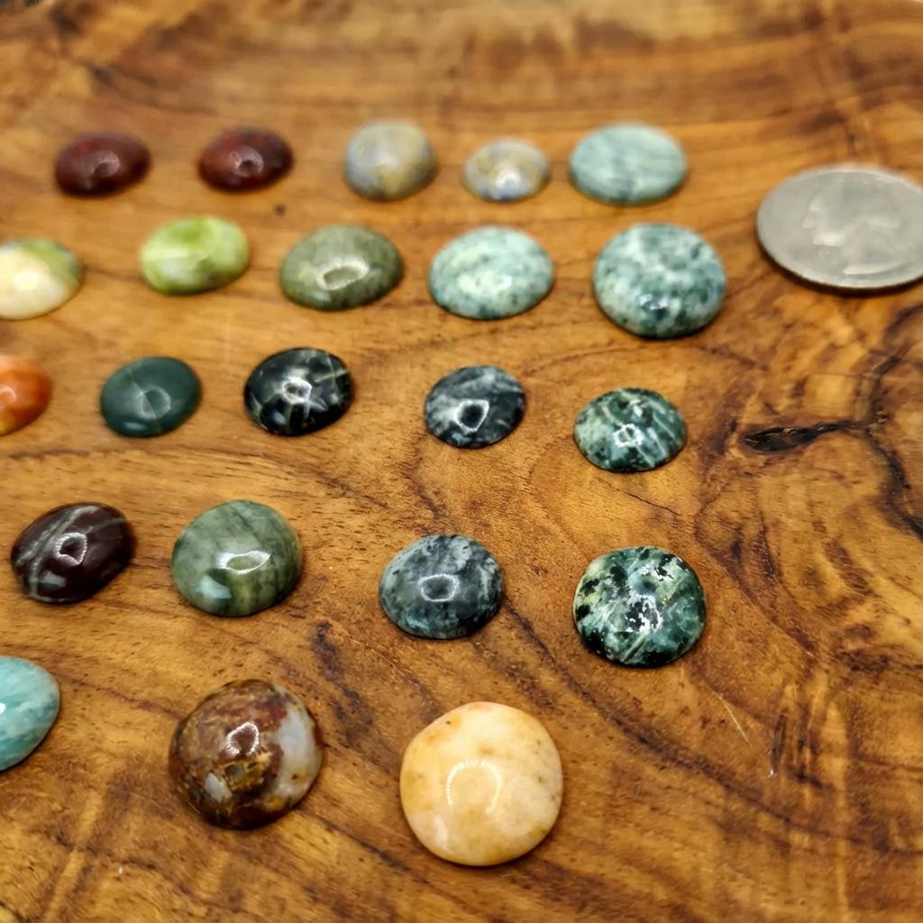Cabochon gemstones displayed on wood with a quarter for size reference. Includes mixed greens, blues, browns, and cream stones with natural veining, mossy textures, and banded chalcedony. Smooth domed shapes reflect studio lighting.