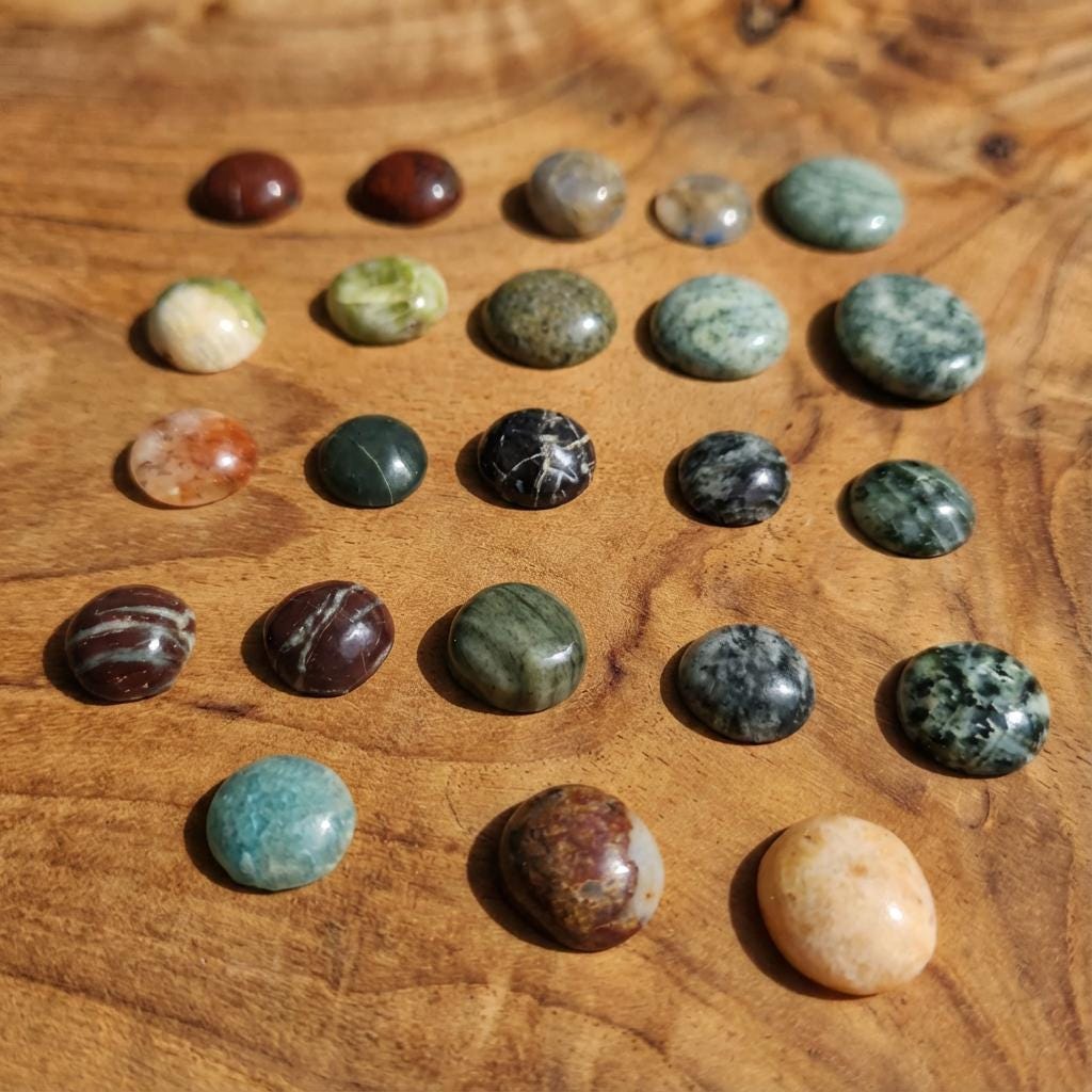 A spread of small polished cabochon gemstones on a warm wood surface, arranged in neat rows. Stones include green jasper, orbicular patterns, red jasper, agate, and mixed chalcedony. Each cabochon has a glossy dome cut, showing natural banding and color variation.