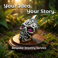 Handcrafted skull pendant with detailed scrollwork and inset pink gemstones, displayed on a wooden surface with a forest backdrop. Represents custom jewelry design services where personal stories become wearable pieces. Bespoke pendants, rings, and heirloom jewelry made through precision CAD and hand finishing.