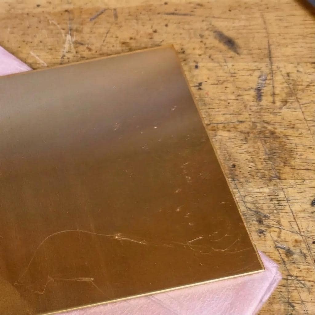 Angled view of a flat brass sheet plate resting on a workshop bench, partially stacked over a protective backing sheet. The brass surface shows a consistent finish with minor scuffs from handling, illustrating real machining stock condition and flatness for CNC routing, engraving, or pocketing operations.