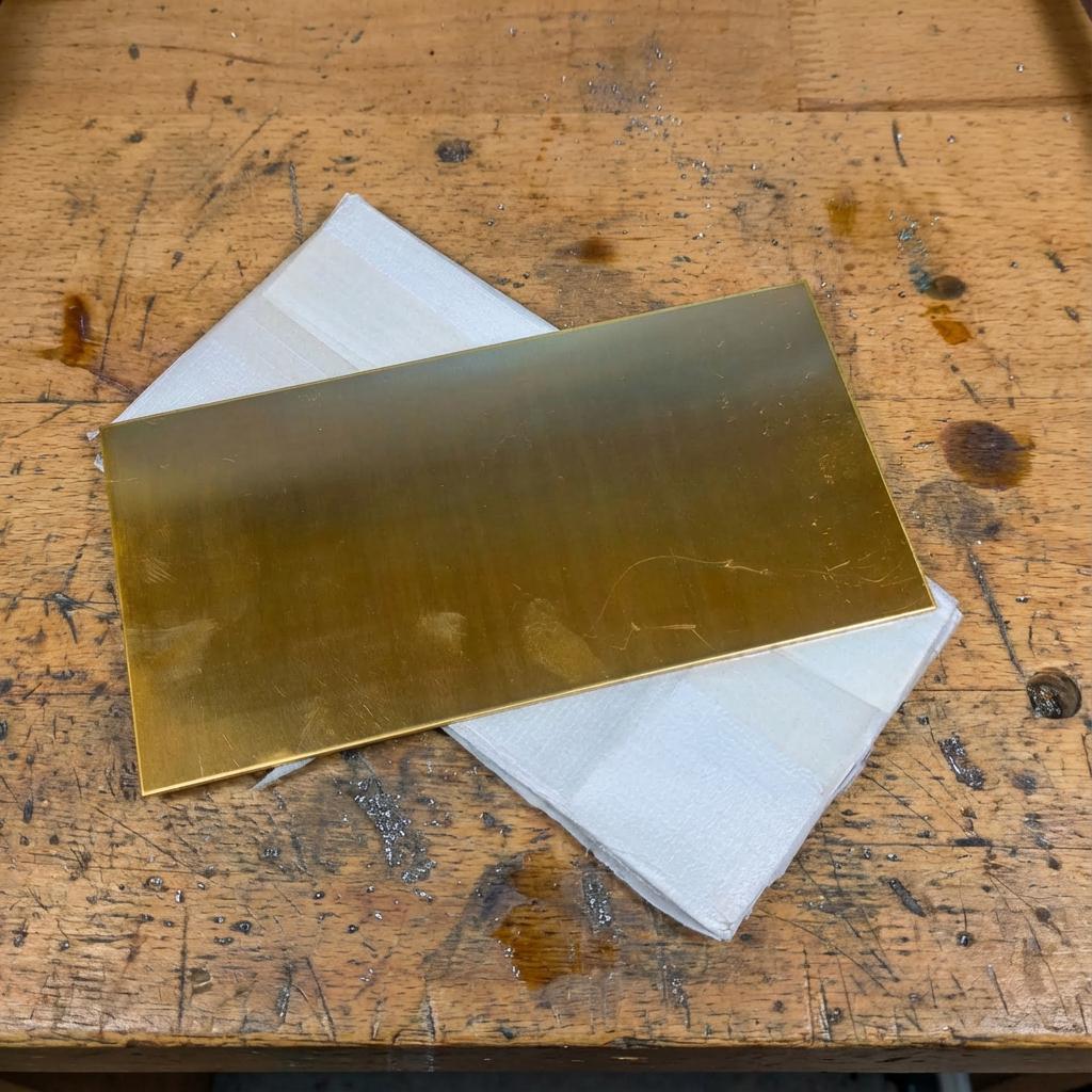 Top-down view of a rectangular brass sheet plate placed over a light-colored backing sheet on a wooden workbench. The image clearly shows overall size, flatness, and proportions of the brass stock, reinforcing its use as CNC machining material rather than decorative or finished metal.