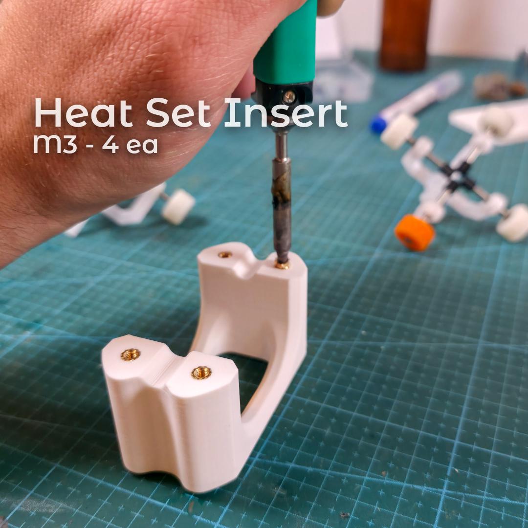 A close-up of a heat-set insert being installed into the Gem-Xfer cure station using a soldering iron. Four M3 brass inserts sit flush in the printed towers. Background includes dop sticks and lapidary tools. Demonstrates hardware installation for users printing the digital STL file and building their own faceting transfer jig with secure threaded anchors.