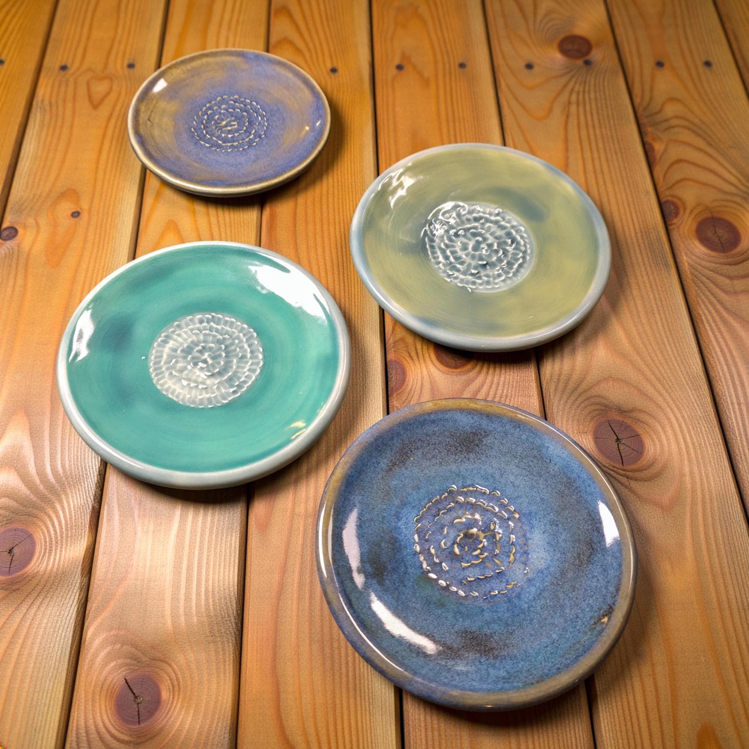 A group of four ceramic plates arranged on a warm wood surface, showcasing glazes in teal, blue, and muted yellow. The plates feature a raised spiral pattern in the center, with glossy finishes that reflect light. The image highlights the organic color blending and handmade character.