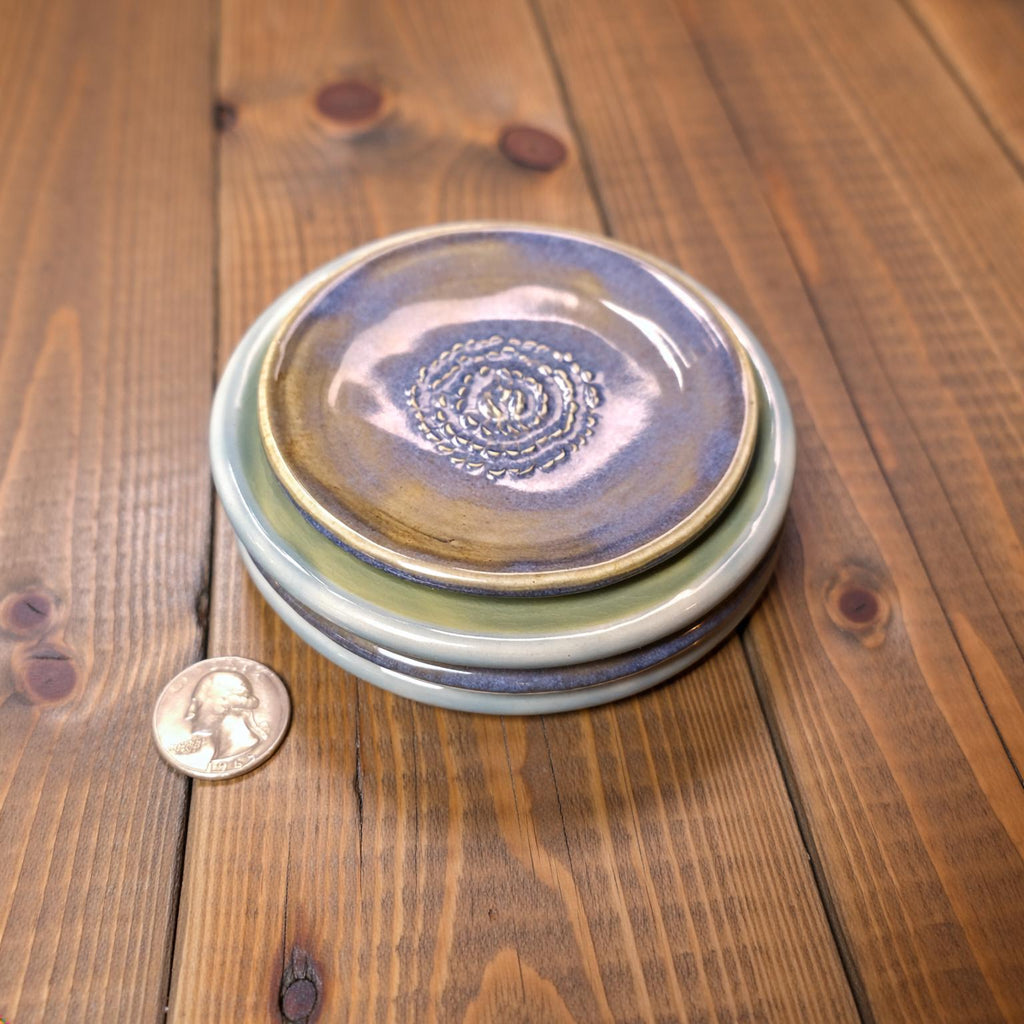 A stack of handmade ceramic plates in layered blue, teal, and yellow glazes sits on a wooden table beside a quarter for scale. The glossy surfaces and textured spiral centers show subtle color transitions, emphasizing the artisan nature of each piece.