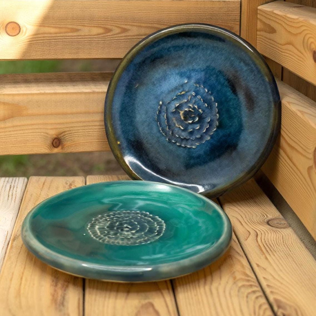 Two ceramic plates displayed on wooden slats outdoors. The top plate is deep blue with a textured spiral center; the bottom plate is teal with similar detailing. Natural light emphasizes the glossy glazes and subtle variations in color.