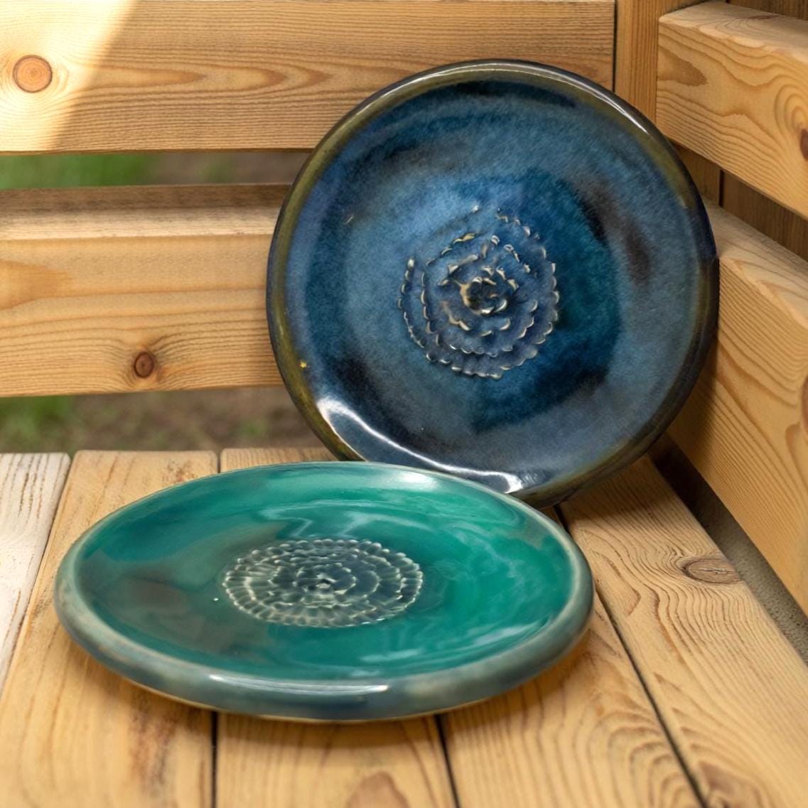 Two ceramic plates displayed on wooden slats outdoors. The top plate is deep blue with a textured spiral center; the bottom plate is teal with similar detailing. Natural light emphasizes the glossy glazes and subtle variations in color.
