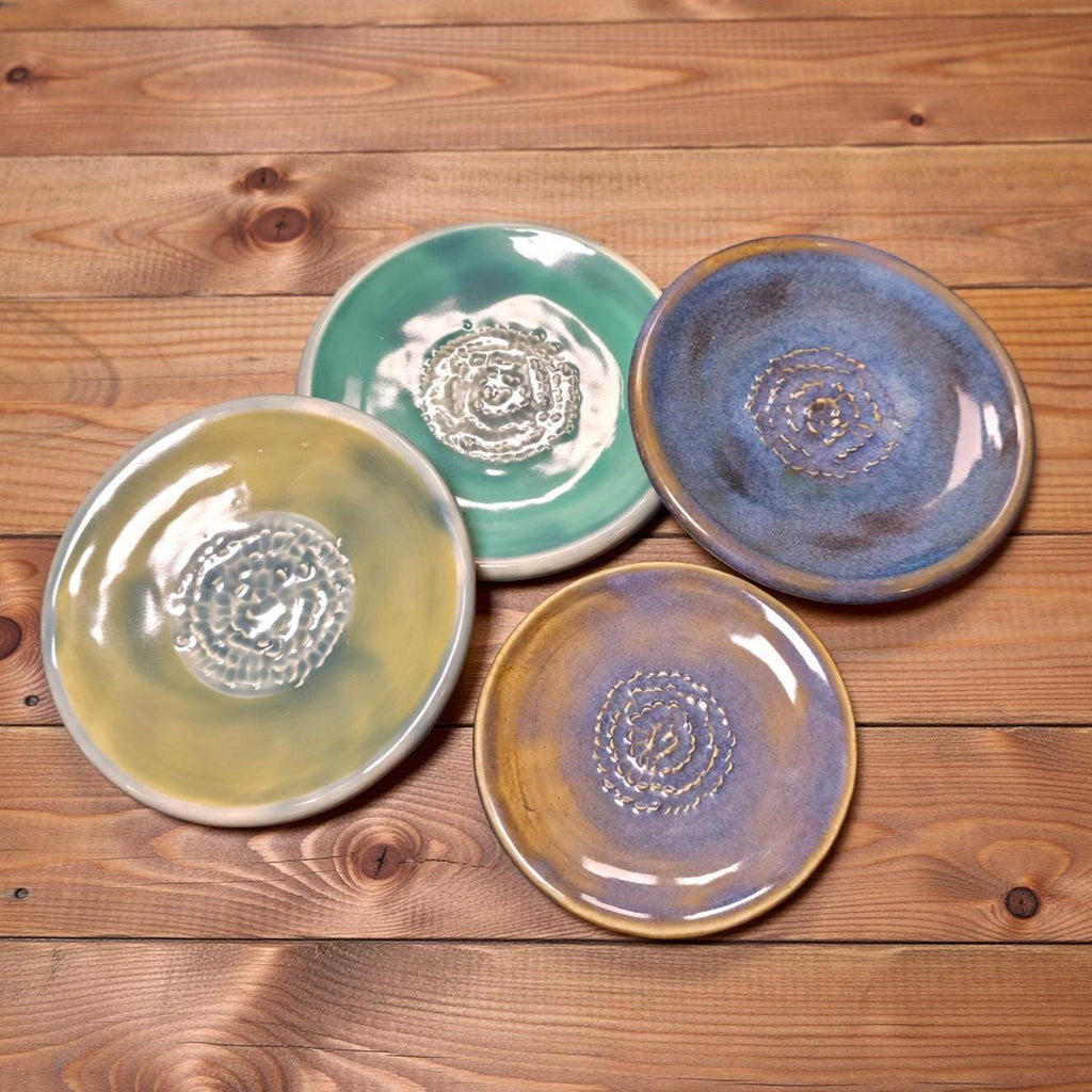 Four handmade ceramic plates displayed on a light wooden tabletop. The plates show varied glazes in teal, blue, and soft yellow with textured spiral centers. Each plate is uniquely finished, emphasizing the artisan craftsmanship and subtle color gradients across the set.