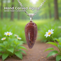 A hand-carved red agate pendant set in a sterling silver bail, hanging from a delicate silver chain against a soft forest background with small white daisies in the foreground. The stone is an elongated teardrop shape with natural reddish and earthy tones. Text on the image reads “Hand Carved Agate – Wabi Sabi Collection,” with a small Aetherium floral mark in the lower right corner.
