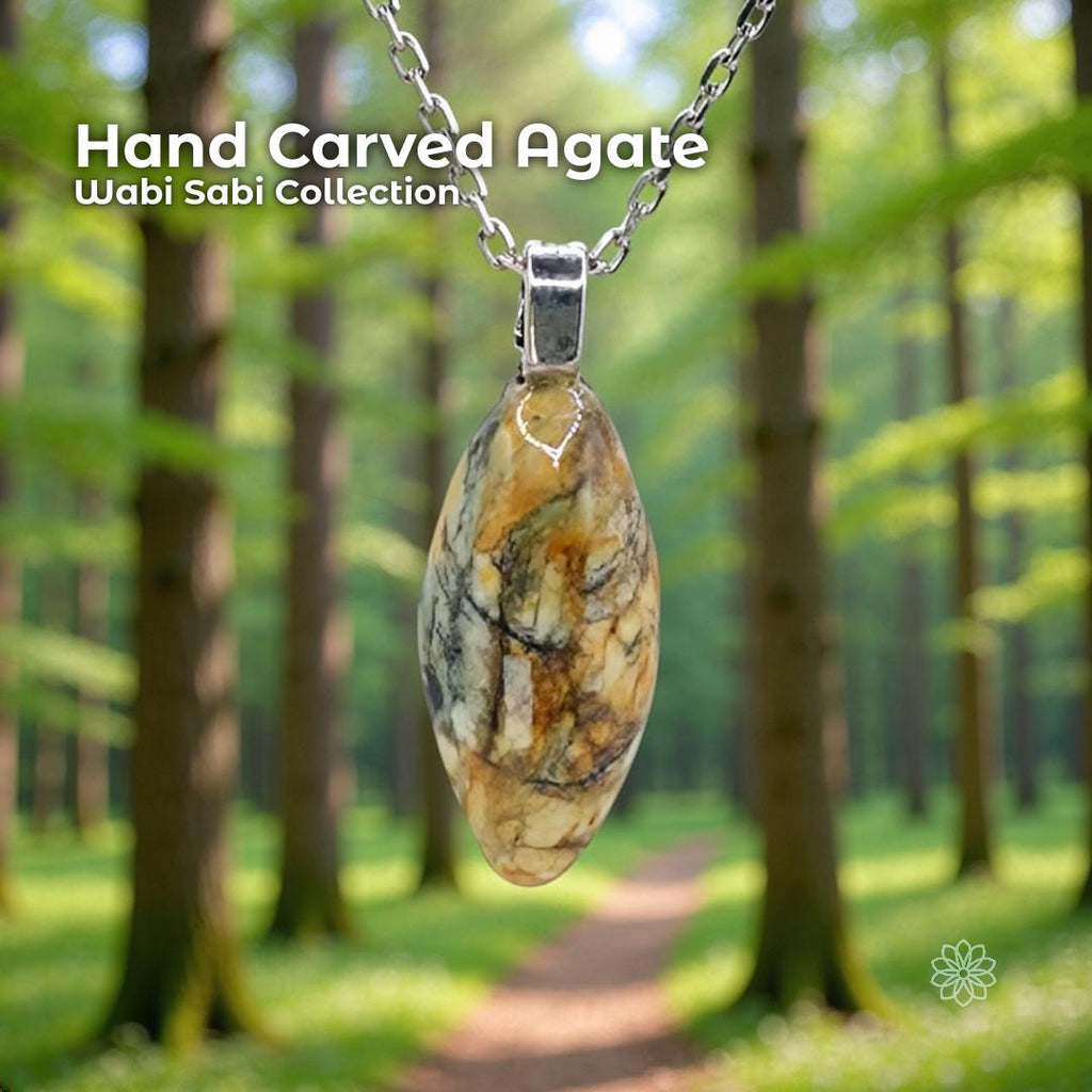 A hand-carved patterned agate pendant set in a sterling silver bail, hanging from a silver chain in a sunlit forest with tall trees and a soft green path. The stone is an elongated oval with natural cream, gold, brown, and gray marbling. Text reads “Hand Carved Agate – Wabi Sabi Collection,” with a small Aetherium floral mark in the lower right corner.