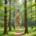 A hand-carved patterned agate pendant set in a sterling silver bail, hanging from a silver chain in a sunlit forest with tall trees and a soft green path. The stone is an elongated oval with natural cream, gold, brown, and gray marbling. Text reads “Hand Carved Agate – Wabi Sabi Collection,” with a small Aetherium floral mark in the lower right corner.