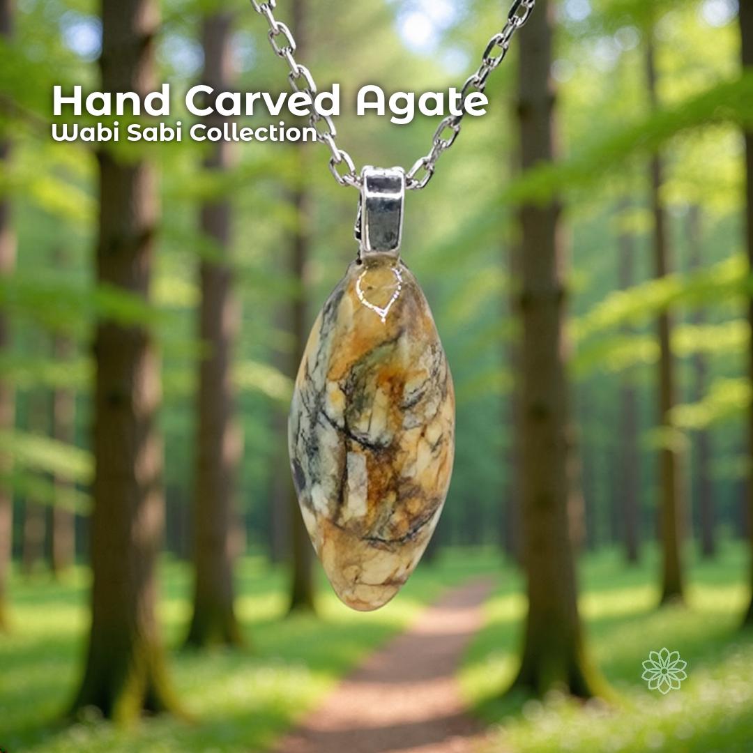 A hand-carved patterned agate pendant set in a sterling silver bail, hanging from a silver chain in a sunlit forest with tall trees and a soft green path. The stone is an elongated oval with natural cream, gold, brown, and gray marbling. Text reads “Hand Carved Agate – Wabi Sabi Collection,” with a small Aetherium floral mark in the lower right corner.