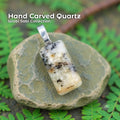A hand-carved dendritic quartz pendant set in a sterling silver bail, resting on a smooth green-gray river stone surrounded by small fern leaves. The quartz is a polished rectangular cabochon with natural white, black, and golden inclusions. Text on the image reads “Hand Carved Quartz – Wabi Sabi Collection,” with a small Aetherium floral mark in the lower right corner.