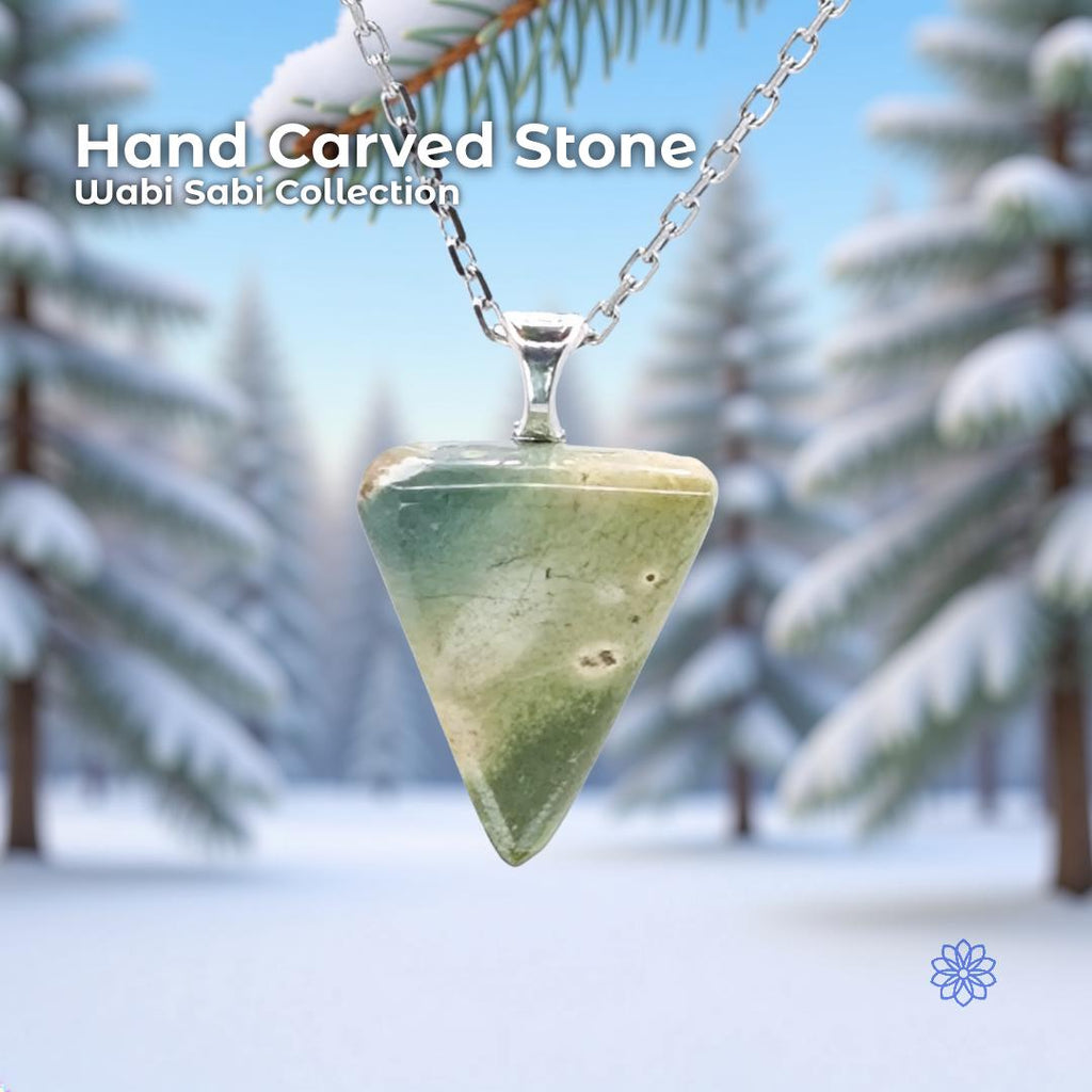 A hand-carved glacial stone pendant set in a sterling silver bail, hanging from a delicate silver chain against a snowy forest background with evergreen branches. The stone is a polished triangular cabochon with soft green, cream, and natural inclusions. Text on the image reads “Hand Carved Stone – Wabi Sabi Collection,” with a small Aetherium floral mark in the lower right corner.