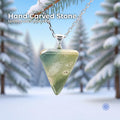 A hand-carved glacial stone pendant set in a sterling silver bail, hanging from a delicate silver chain against a snowy forest background with evergreen branches. The stone is a polished triangular cabochon with soft green, cream, and natural inclusions. Text on the image reads “Hand Carved Stone – Wabi Sabi Collection,” with a small Aetherium floral mark in the lower right corner.