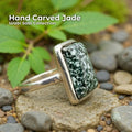 Hand carved nephrite jade ring featuring a dark green stone with white speckled patterns, set in a solid sterling silver band. The ring rests on smooth river stones and sand with green leaves in the background, creating an earthy Pacific Northwest aesthetic and emphasizing the handmade, nature-inspired design.