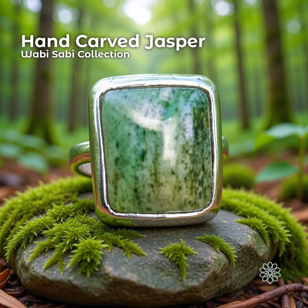 Hand carved green jasper cabochon ring set in a solid sterling silver band. The stone has vertical bands of soft and deep green with a raw, unsealed finish. The ring sits on a moss-covered rock in a lush forest setting, surrounded by soft green light and blurred trees, emphasizing its nature-inspired Pacific Northwest style.
