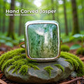 Hand carved green jasper cabochon ring set in a solid sterling silver band. The stone has vertical bands of soft and deep green with a raw, unsealed finish. The ring sits on a moss-covered rock in a lush forest setting, surrounded by soft green light and blurred trees, emphasizing its nature-inspired Pacific Northwest style.