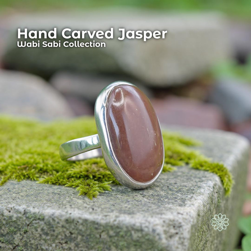 A sterling silver ring featuring an oval red jasper cabochon with soft brown-red tones, set in a smooth hammer-set bezel. The ring sits on bright green moss and stone, photographed outdoors to emphasize its natural Pacific Northwest style, raw unsealed stone, and grounding, nature-aligned aesthetic.