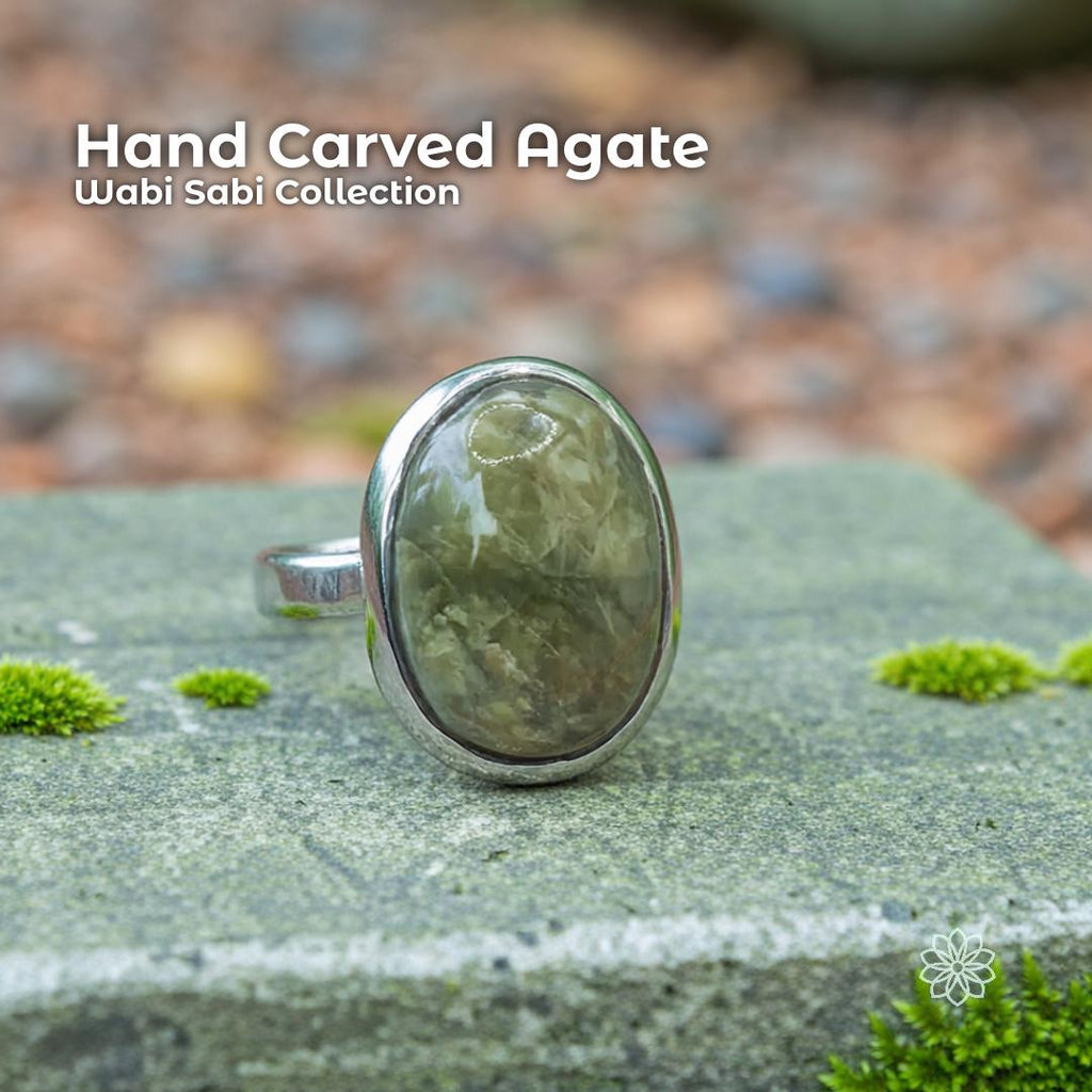 A handcrafted sterling silver ring featuring an oval olive agate cabochon with soft green and earthy inclusions, displayed outdoors on a mossy stone. The polished natural gemstone sits in a hammer-set bezel, surrounded by forest textures and muted tones that emphasize its Pacific Northwest, nature-inspired style.