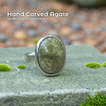 A handcrafted sterling silver ring featuring an oval olive agate cabochon with soft green and earthy inclusions, displayed outdoors on a mossy stone. The polished natural gemstone sits in a hammer-set bezel, surrounded by forest textures and muted tones that emphasize its Pacific Northwest, nature-inspired style.