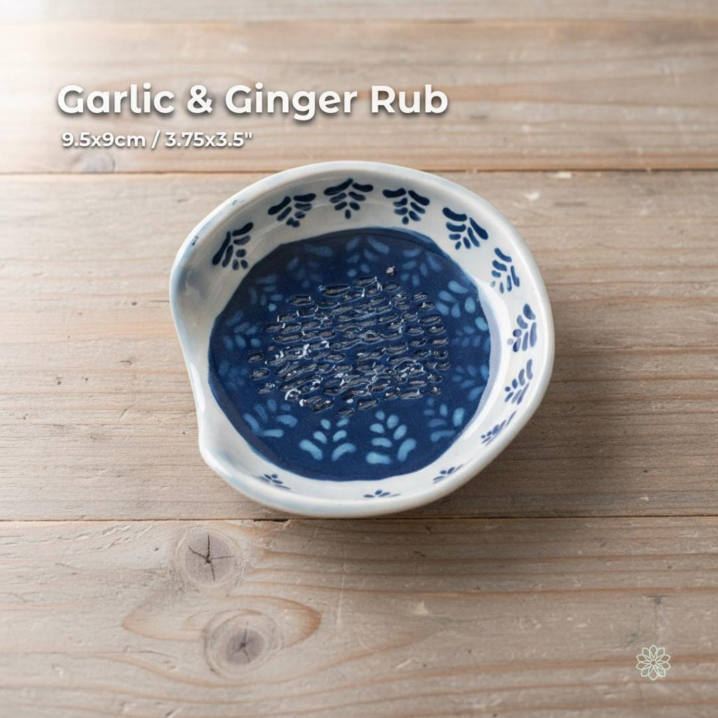 A small handmade ceramic garlic and ginger grater dish featuring carved ridges in the center, surrounded by a deep cobalt glaze with hand-painted botanical blue-and-white patterns around the rim. Displayed on a rustic wooden surface. Functional for cooking prep or as a decorative kitchen dish. Part of the Aetherium × Archibald artisan pottery series.