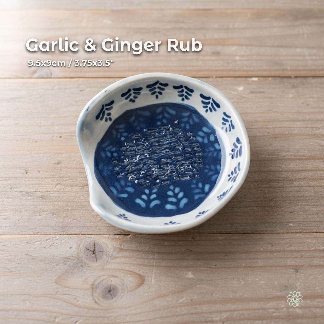 A small handmade ceramic garlic and ginger grater dish featuring carved ridges in the center, surrounded by a deep cobalt glaze with hand-painted botanical blue-and-white patterns around the rim. Displayed on a rustic wooden surface. Functional for cooking prep or as a decorative kitchen dish. Part of the Aetherium × Archibald artisan pottery series.
