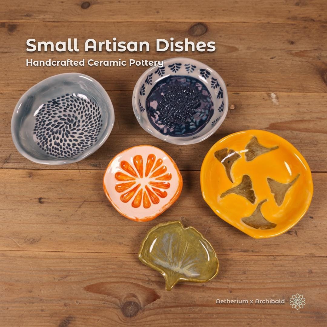 A curated collection of five handmade artisan ceramic dishes displayed on a warm wooden tabletop. Designs include a gray-blue spotted dish, a navy botanical garlic rub, an orange slice, a yellow chanterelle mushroom dish, and a green ginkgo leaf dish. Each piece shows unique glazing, hand-carved details, and organic shapes. Presented as the Aetherium × Archibald Small Artisan Dish Collection.