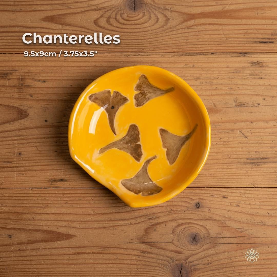 A round handmade ceramic dish in warm golden yellow glaze featuring carved chanterelle mushroom motifs in earthy brown tones. Displayed on a rustic wooden table, the dish highlights its shallow form and glossy surface. Suitable as a trinket dish, jewelry holder, kitchen accent, or small serving piece. From the Aetherium × Archibald artisan pottery series.