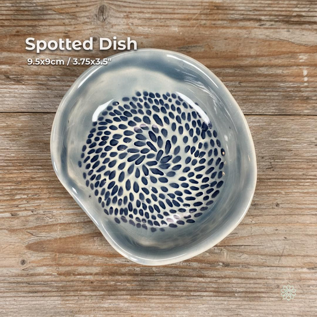 A handmade ceramic dish with an irregular organic shape, glazed in soft gray-blue with a dense pattern of hand-painted navy spots swirling from center to rim. Shown on a textured wooden surface under natural light. Usable as a catchall dish, jewelry bowl, kitchen accent, or incense holder. Part of the Aetherium × Archibald handcrafted pottery line.