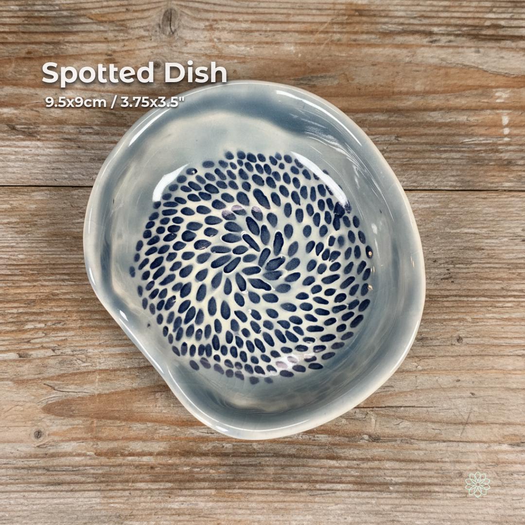 A handmade ceramic dish with an irregular organic shape, glazed in soft gray-blue with a dense pattern of hand-painted navy spots swirling from center to rim. Shown on a textured wooden surface under natural light. Usable as a catchall dish, jewelry bowl, kitchen accent, or incense holder. Part of the Aetherium × Archibald handcrafted pottery line.