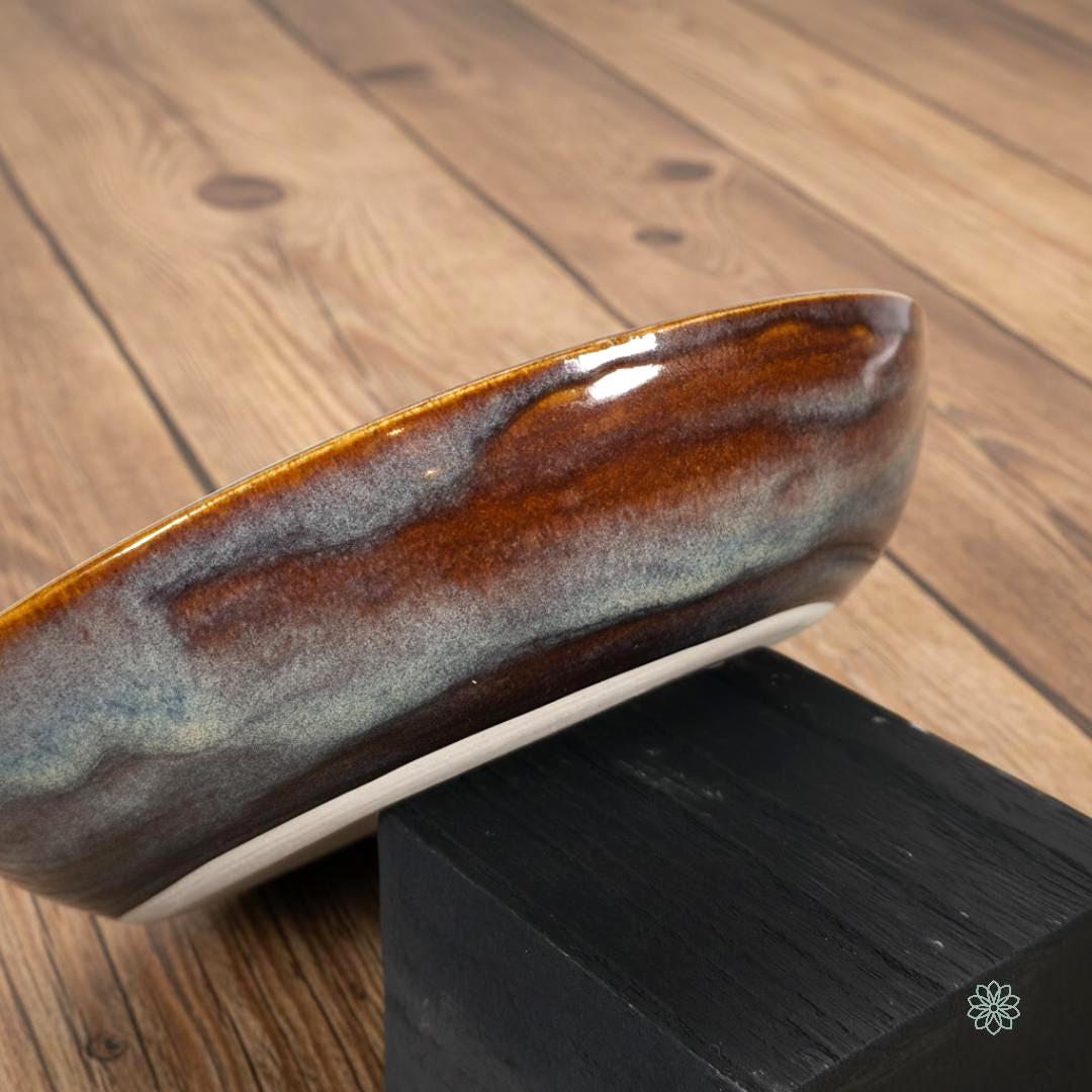 “Side profile of a handmade stoneware pasta bowl featuring a reactive glaze that fades from deep amber to blue and gray tones. The curved walls and smooth rim show the bowl’s depth and craftsmanship. Displayed on a wood backdrop as part of the Aetherium × Archibald pottery line.”