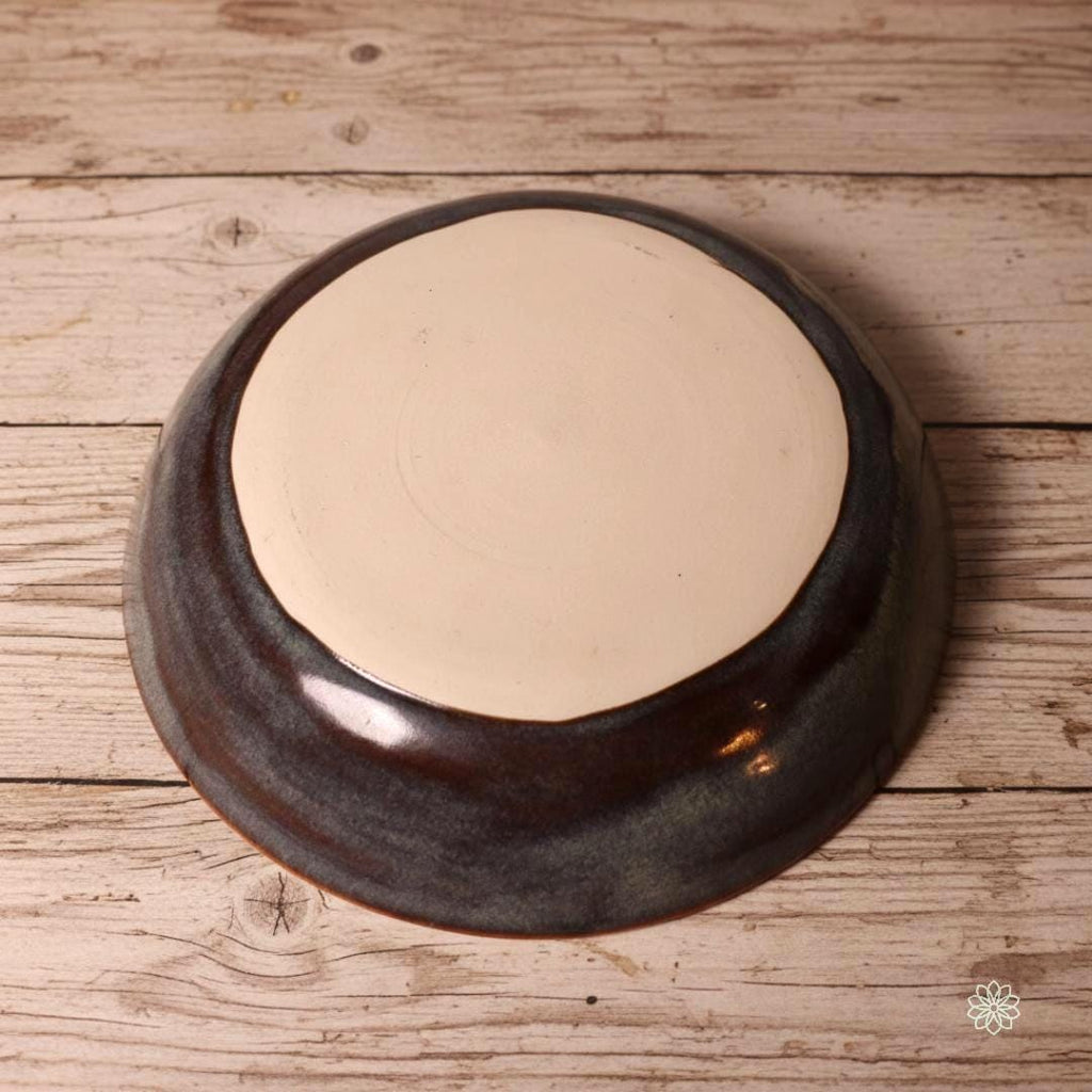 “Underside of a handmade stoneware pasta bowl, showing the unglazed clay base and natural wheel-thrown texture. The reactive glaze wraps around the sides with blue, gray, and rust tones. Displayed on a rustic wood table as part of the Aetherium × Archibald collection.”