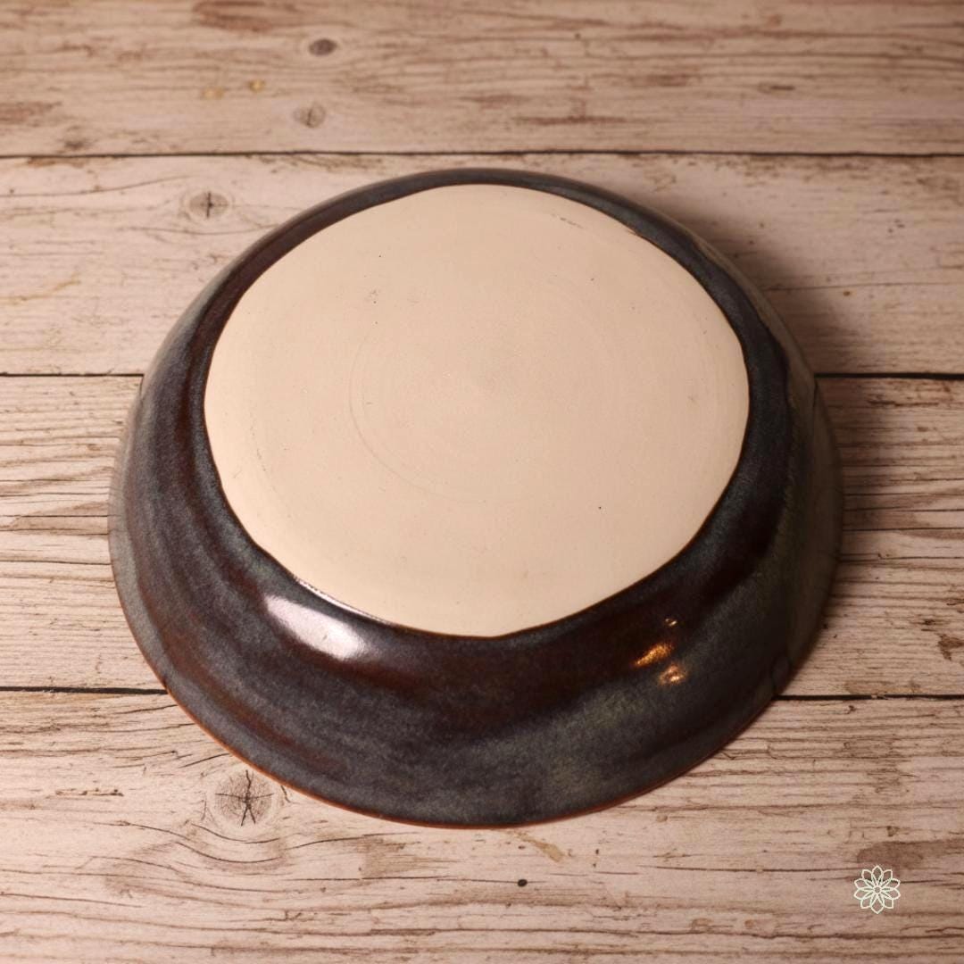 “Underside of a handmade stoneware pasta bowl, showing the unglazed clay base and natural wheel-thrown texture. The reactive glaze wraps around the sides with blue, gray, and rust tones. Displayed on a rustic wood table as part of the Aetherium × Archibald collection.”