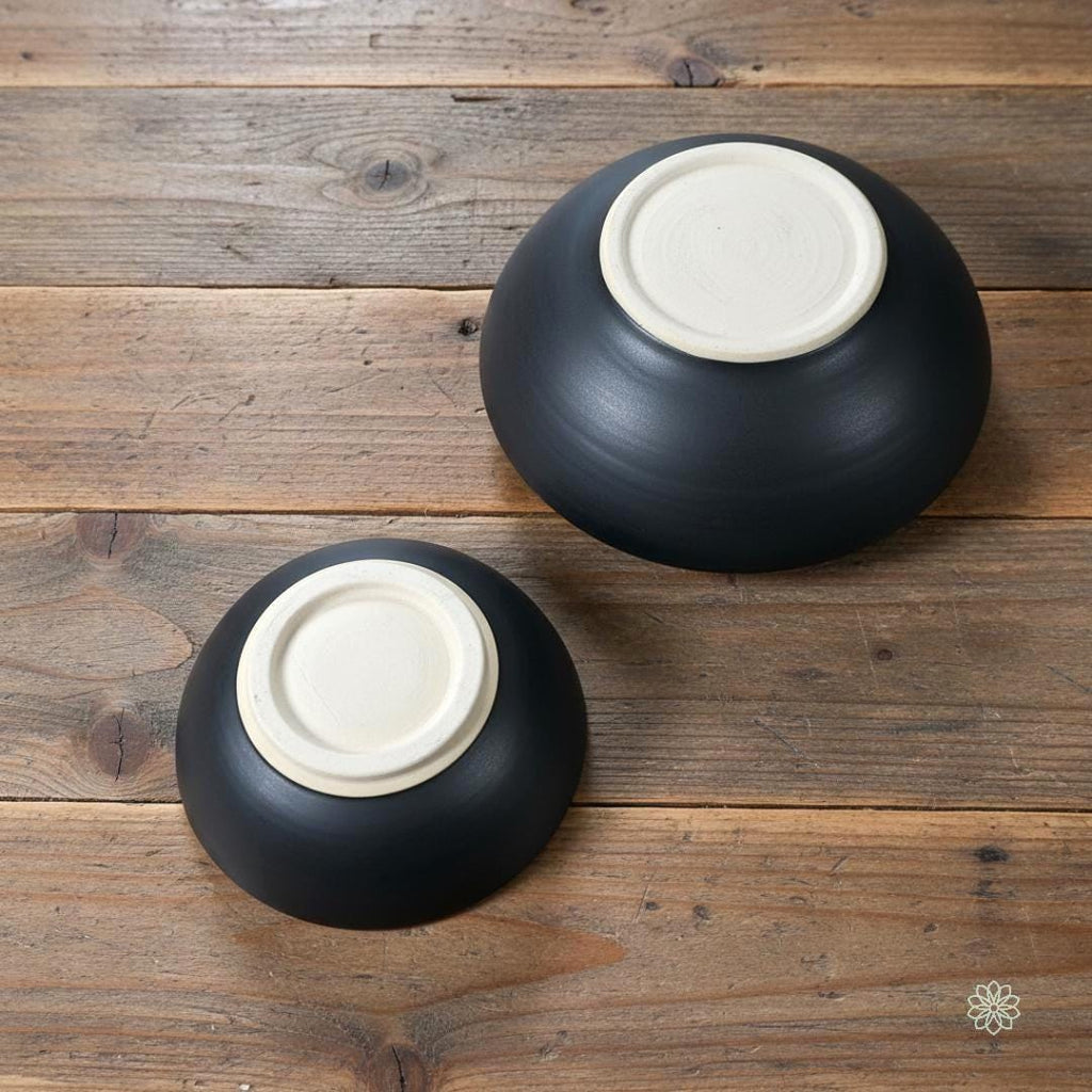 “Underside view of two handmade stoneware bowls showing smooth unglazed bases and matte black exteriors. The foot rings and clay texture are visible, emphasizing craftsmanship. Displayed on a wood tabletop.”