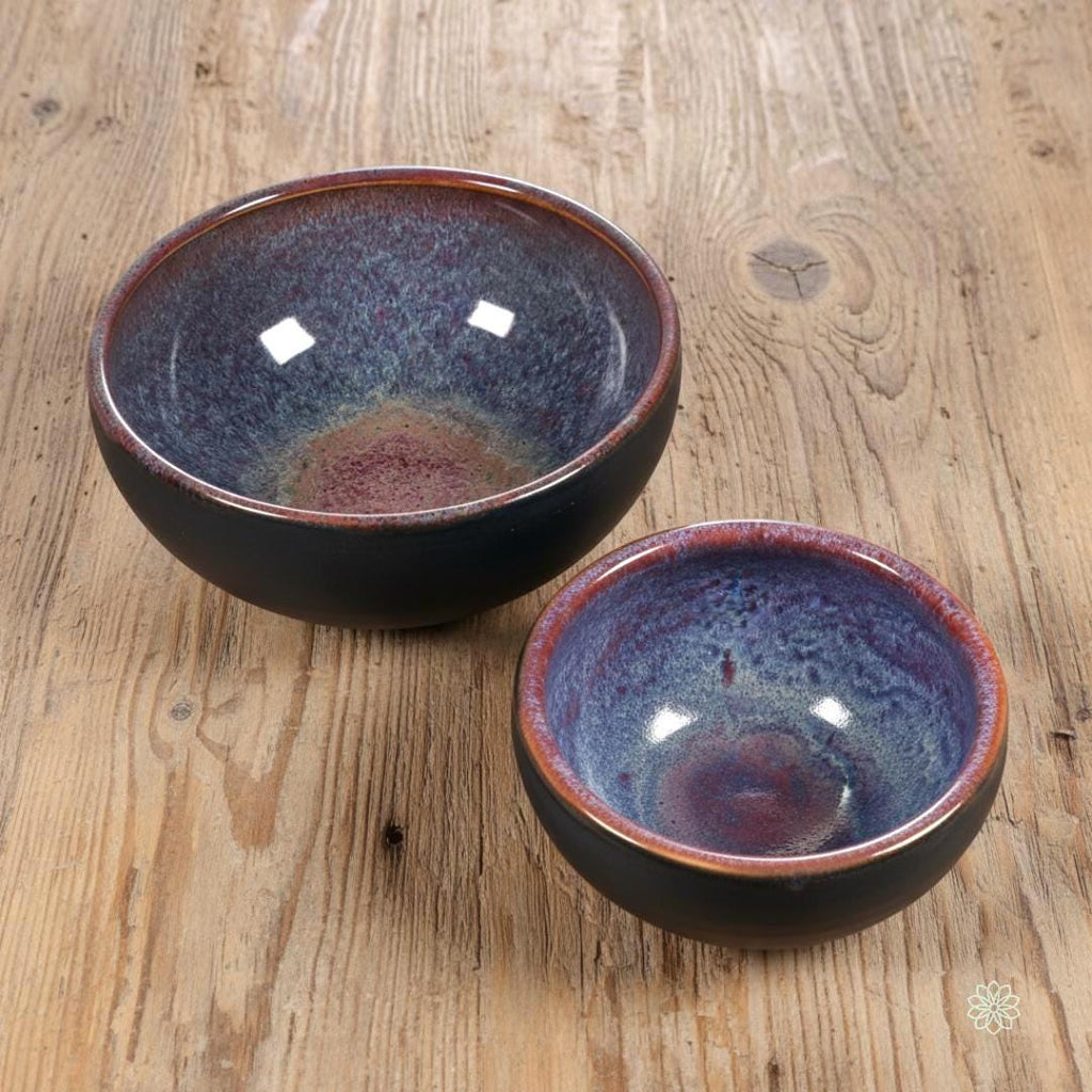 “Two handcrafted stoneware bowls with matching reactive glaze interiors in violet, blue, and red tones. Both bowls face upward to show the layered crystalline patterns. Matte black exteriors and glossy interiors on a wood tabletop.”