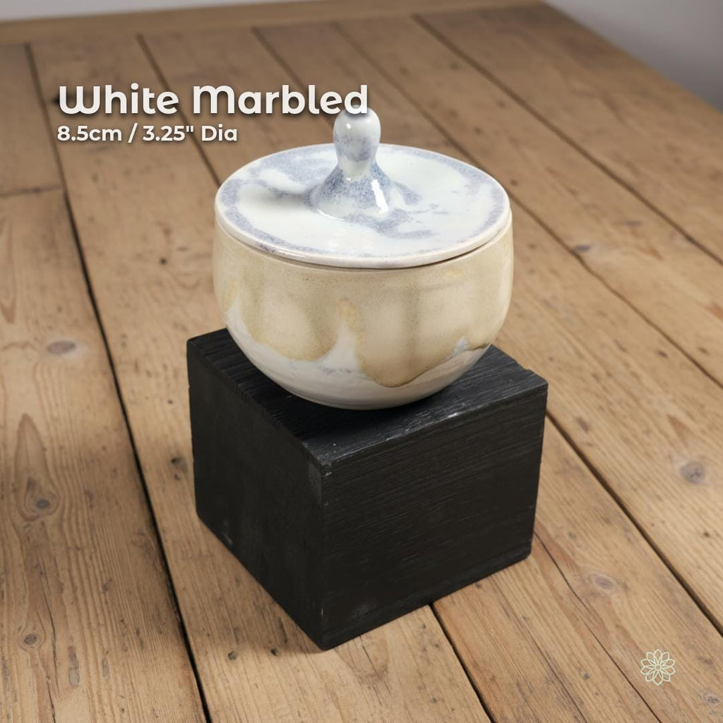 “White marbled ceramic altar jar resting on a black block. On-image text reads ‘White Marbled’ and ‘8.5cm / 3.25" Dia.’ Smooth rounded lid with sculpted handle. Displayed on light wood surface.”