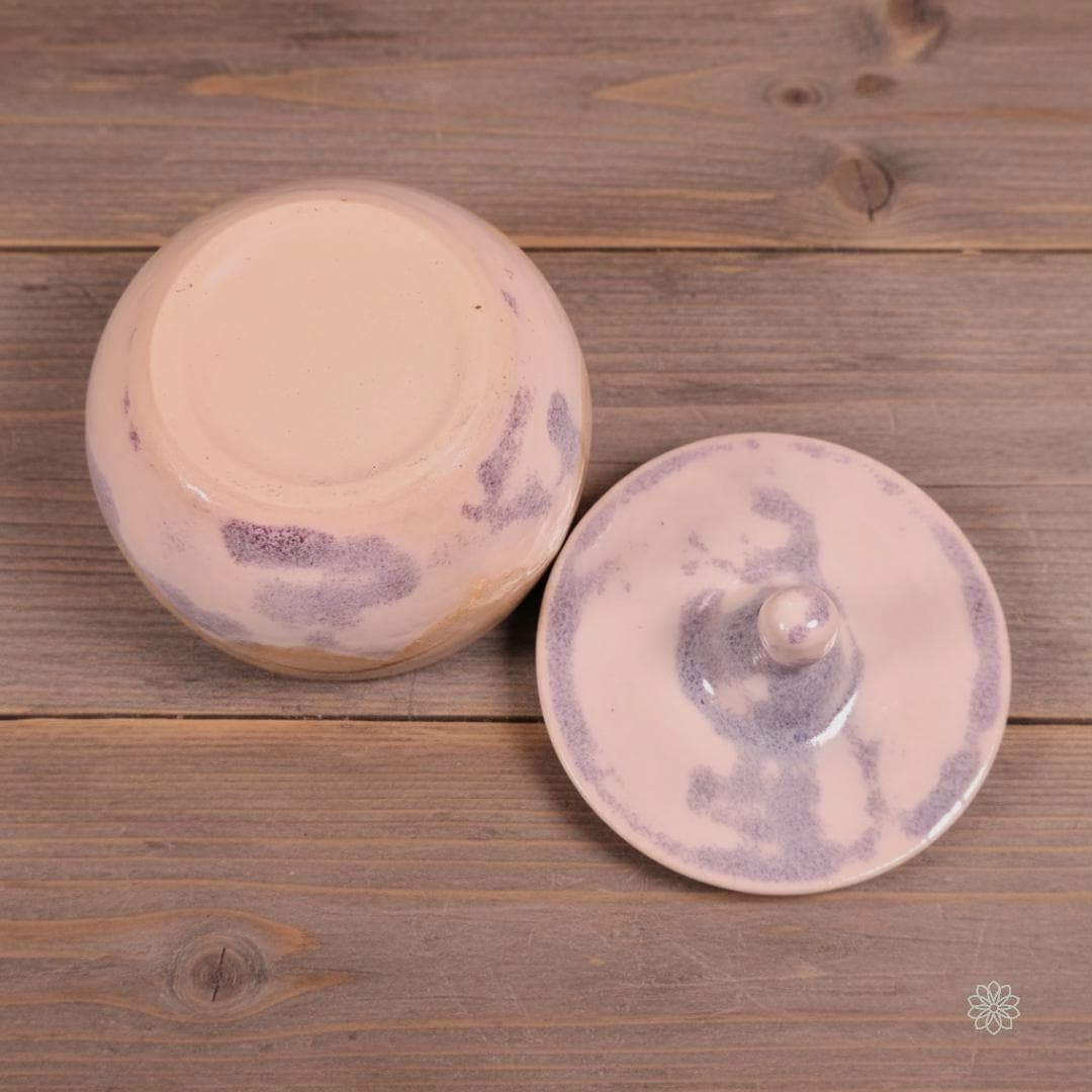 Underside details of the White Marbled altar jar. The jar is flipped to show its unglazed foot ring and subtle lavender-blue marbling on the exterior. The lid sits beside it, facing upward to reveal its marbled glaze pattern and rounded handle. Photographed on warm plank wood for continuity.