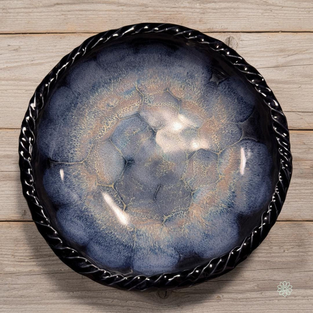 Top-down view of a large braided-rim reactive glaze stoneware bowl on a pale wood surface. The interior glaze shows concentric blue crystalline patterns with soft transitions from dark cobalt near the rim to lighter blue and tan toward the center. The bowl is presented as a handmade serving or display piece.