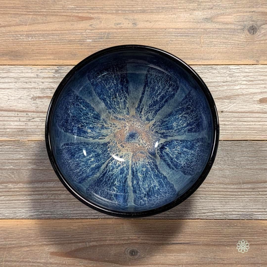 Top-down view of a smaller reactive glaze stoneware bowl with a deep radial crystalline pattern. Dark blue lines radiate from a warm tan center, creating a starburst effect inside the glossy bowl. The bowl sits on a weathered wooden table with mixed light and dark planks.