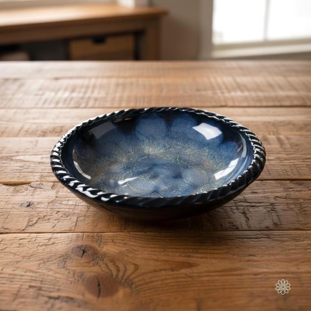 Large braided-rim reactive glaze stoneware bowl centered on a rustic wood table in front of a softly lit window. The interior shows layered blue crystalline patterns with subtle tan tones and a glossy finish. The bowl is shown as a functional serving or display piece for the home.