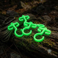 Four glow-in-the-dark bioplastic paracord hooks resting on a log with paracord and a carabiner nearby. Nighttime outdoor scene emphasizes high visibility, quick gear access, and campsite organization for camping and survival setups.