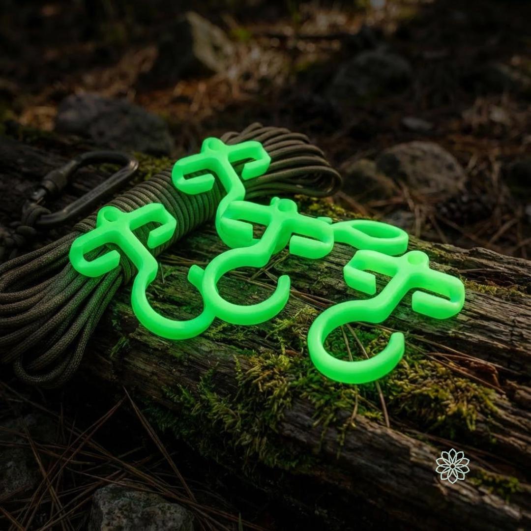Four glow-in-the-dark bioplastic paracord hooks resting on a log with paracord and a carabiner nearby. Nighttime outdoor scene emphasizes high visibility, quick gear access, and campsite organization for camping and survival setups.