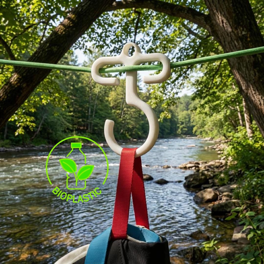 Full-size bioplastic paracord hook hanging on a green ridgeline above a river in a forest. The hook holds camping gear straps and shows the two-in-one loop-and-hook design. Photo highlights outdoor use, durability, and lightweight camp organization for backpackers and campers.