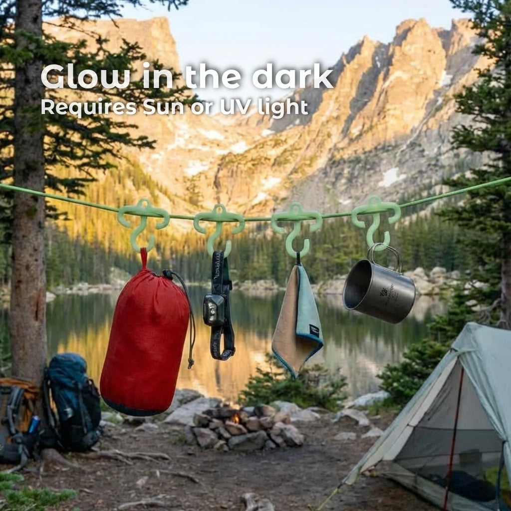 Row of mini paracord hooks glowing green at dusk, clipped to a taut line holding a red stuff sack, headlamp, microfiber towel, and metal mug. Background shows a mountain lake campsite. Built for ultralight backpacking and quick campsite organization.