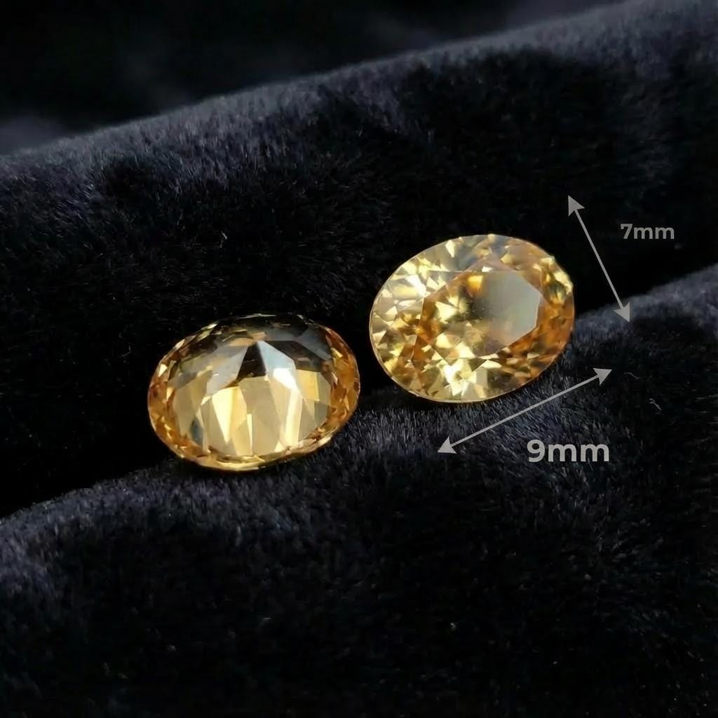 “Two champagne oval cubic zirconia gemstones resting on black velvet. One stone is face-up and the other shows the pavilion facets. Overlay text indicates 9×7 mm dimensions. The gems display warm gold tones and precise faceting for jewelry making or custom bridal designs.”