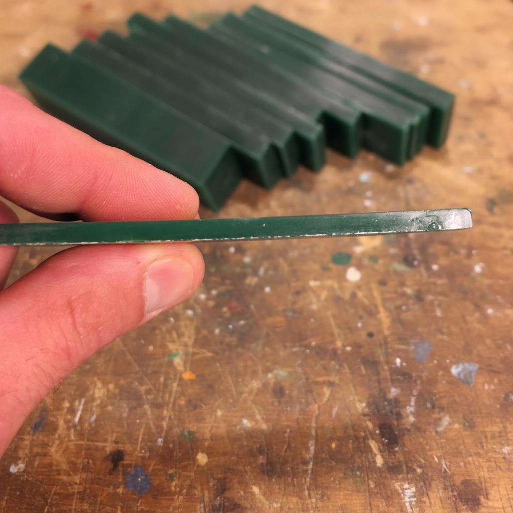 “A thin green carving-wax strip held between fingers, showing a clean machined edge and smooth surface. Several wax slabs are stacked in the background on a jeweler’s workbench. This type of machinable wax is used for lost-wax casting, CNC engraving, ring carving, and fine jewelry model making.”