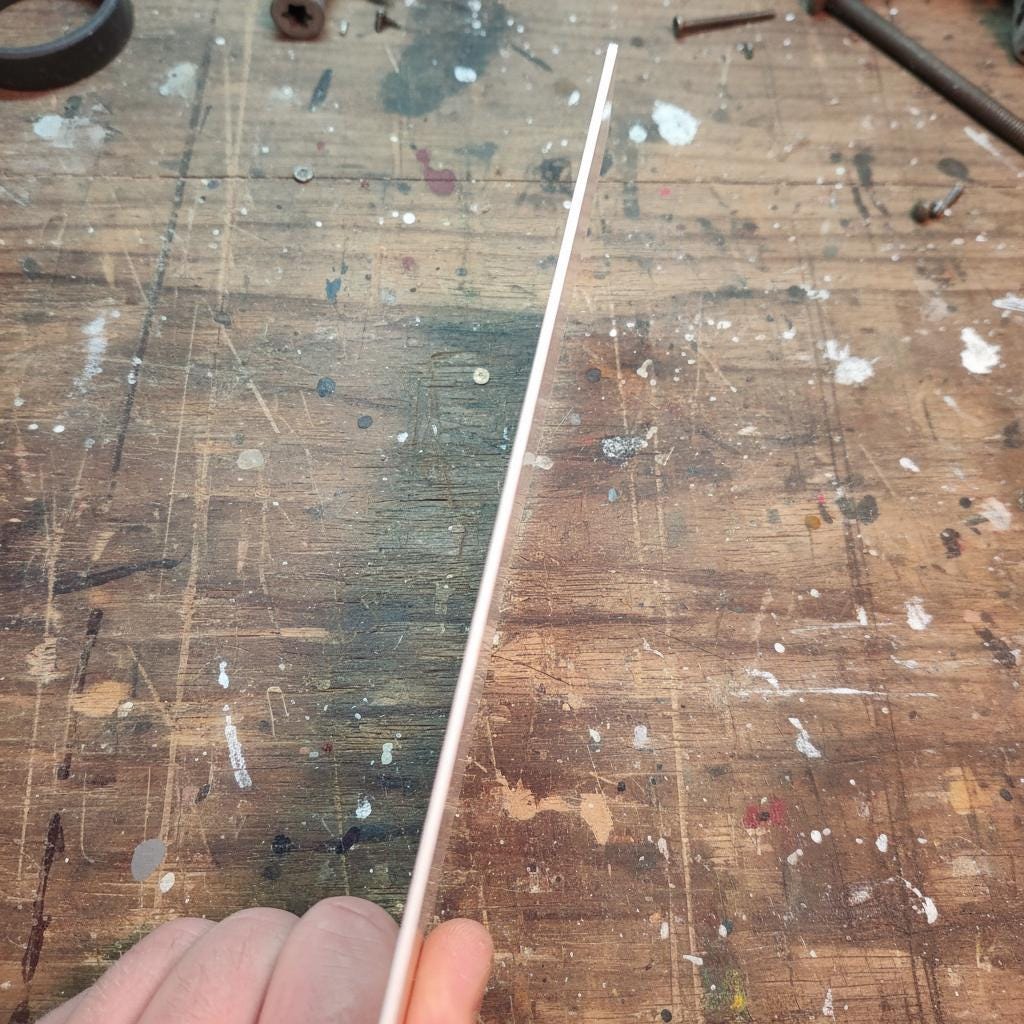 “A thin double-sided copper-clad PCB sheet held on edge to show its consistent thickness and fiberglass core. Both sides are laminated with copper, making it suitable for CNC isolation routing, chemical etching, RF prototyping, and custom two-layer circuit board fabrication. A worn workbench is visible behind it.”