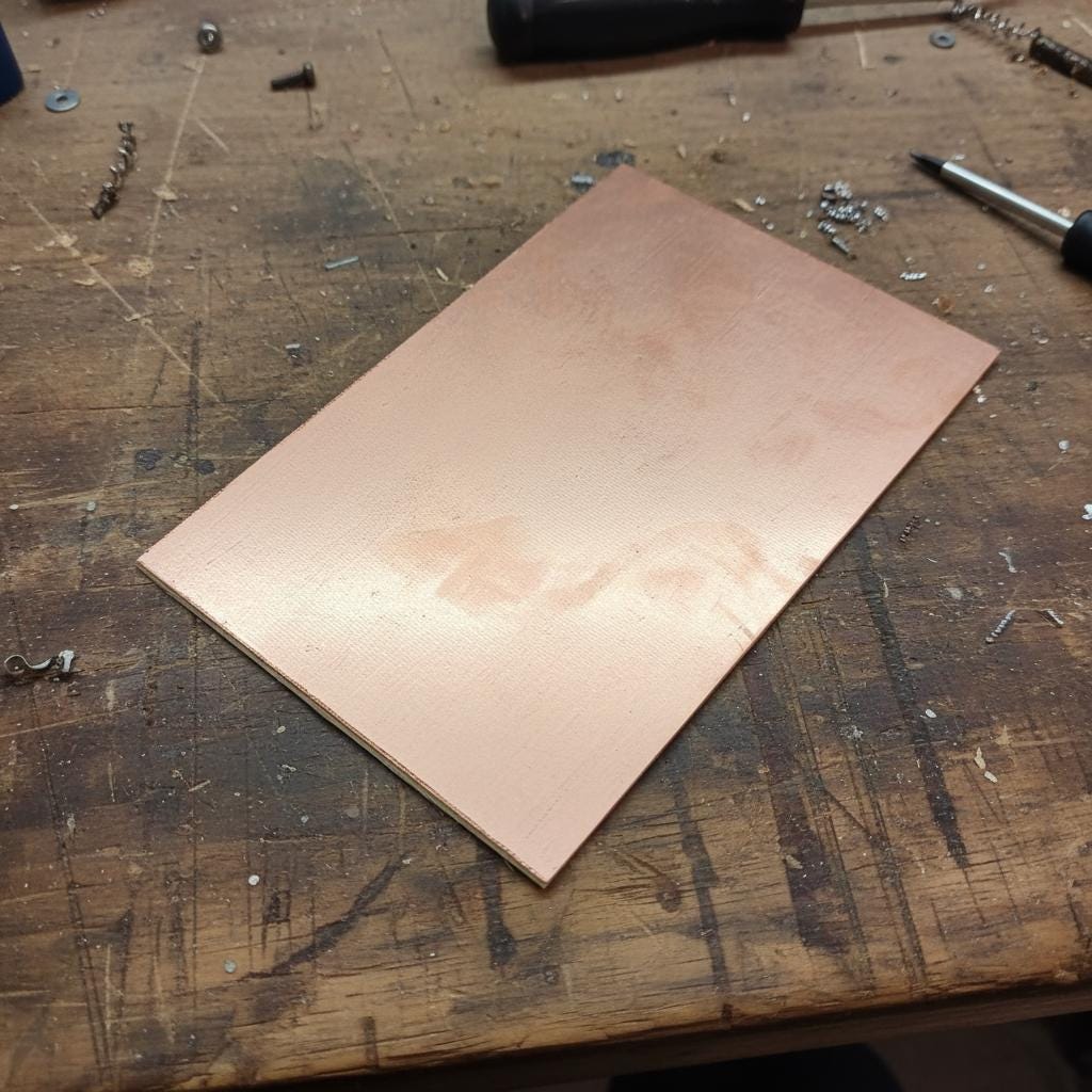 “A large double-sided copper-clad PCB blank resting on a workshop table with a soldering iron, pliers, and scattered components nearby. The board features copper on both sides, ideal for etching or CNC routing dual-layer circuits, PCB prototyping, and electronics fabrication.”