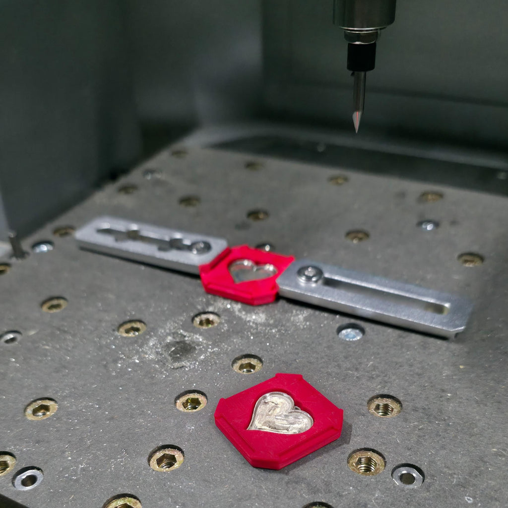 “Close-up of a desktop CNC milling machine bed showing a precision engraving setup for custom jewelry. A small carbide bit hangs above a metal fixture holding a red 3D-printed jig with a heart-shaped silver pendant blank, with another matching jig and heart on the perforated work table, illustrating prototype machining for personalized jewelry parts.”